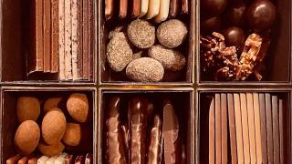 A close-up of an assorted chocolate box divided into six compartments, each holding multiple different types of chocolates: truffles, chocolate-dipped sticks, caramel clusters, and layered bars.
