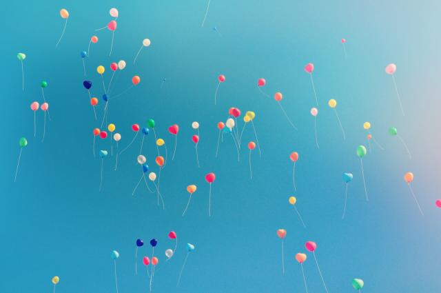 Dozens of small, brightly coloured balloons drifting upward against a clear blue sky.