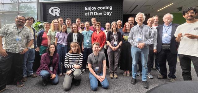 About two dozen R Dev Day participants gathered for a group photo in a bright, glass-walled atrium, in front of a sign reading 'Enjoy coding at R Dev Day'.