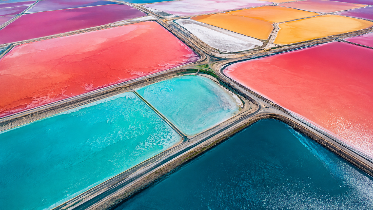 An aerial-style view of broad salt ponds in pink, coral, orange, turquoise, and deep blue, divided by narrow roads.